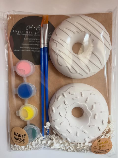 A paint-your-own donut kit: two white plaster donuts, four pots of paint, and a brush in a clear bag with the Absolute Creations card.