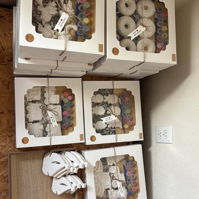 Stacks of white gift-box kits tied with twine, each with plaster pieces, a strip of paints, and brushes.